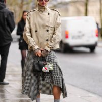 Guest wears dark brown sunglasses, gold earrings, light brown high neck double breasted trench jacket, gold bracelets, gold rings, white black...