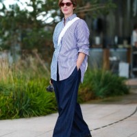 Guest wears dark brown sunglasses, light blue white lined button up long sleeve shirt, silver bracelet, white leather bag, navy blue denim jean...
