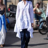 Guest wears dark brown sunglasses, white Comme Des Garcons button up long sleeve shirt dress with ruffled layered bottom, silver rings, navy dark...