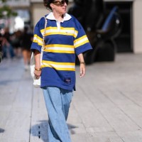 Guest wears dark brown sunglasses, white navy blue yellow striped short sleeve oversized shirt, white leather bag, a gold bracelet, a watch, gold...