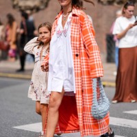 Guest wears dark brown sunglasses, white necklace, bright orange and white checkered pattern trench coat, white mini dress with large collar and...