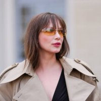Guest wears dark orange sunglasses, black deep v-neck shirt, light brown trench coat, outside Tod’s, during the Milan Fashion week Women's...
