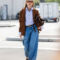 Guest wears denim wide leg jeans, brown jacket, bag, blue button shirt outside Prada during the Milan Fashion Week Menswear Spring/Summer 2025 on...