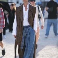 Guest wears glasses, a brown sleeveless long checked trench coat, a white shirt, wide leg blue denim jeans, outside Casablanca, during the Womenswear...