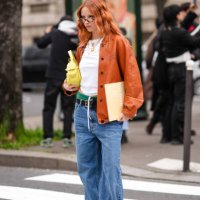 Guest wears glasses, an orange leather jacket , a white cropped top, green underwear, a belt, blue wide-leg denim pants / jeans , a yellow leather...