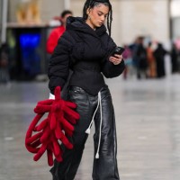 Guest wears gold earrings, black tailored puffer jacket, shiny black wide leg leather pants, dark red 3D graphic bag, white sneaker shoes, outside...