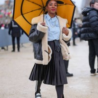 Guest wears gold earrings, gold necklace, light blue lined buttoned up long sleeve shirt, black pleated midi skirt, shiny black leather gloves, shiny...