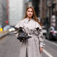 Guest wears gold earrings, light gray Altuzarra belted trench coat, shiny black large bronzed studs design Altuzarra origami leather bag, outside...