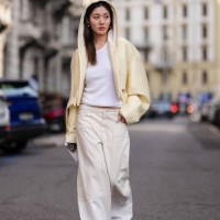 Guest wears gold earrings, light pale yellow hooded cropped leather jacket, white tank top, cream low-rise baggy denim jean pants, outside Sportmax,...