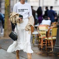Guest wears gold earrings, white oversized t-shirt with black graphic print, light brown beige cropped trench coat, white sheer mesh lace midi skirt,...