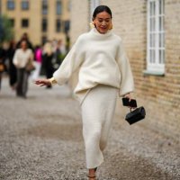 Guest wears gold large earrings, a white latte wool turtleneck pullover, a white latte wool long skirt, a black shiny leather handbag, black shiny...