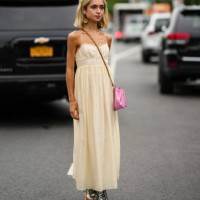 Guest wears gold pendant earrings, a beige square neck long dress, a pale pink shiny leather crossbody bag, silver shiny leather laces ankle boots,...