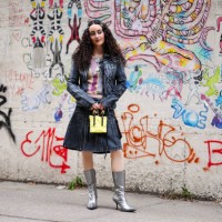 Guest wears hair pins with pearl details, a silver necklace, a grey and purple crop top with prints and yellow striped details / tie and dye, a black...
