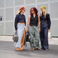 Guest wears light brown fuzzy hat, black sunglasses, black long sleeve cropped shirt with a Chanel pendant, light brown maxi skirt, light gray white...