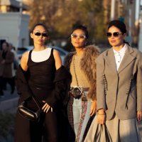 Guest wears long black skirt, white top, black top and black coat and a guest wears wide jeans, beige top and fur jacket and a guest wears grey long...