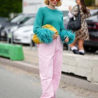 Guest wears orange aviator sunglasses, a gold necklace, a green braided wool with embroidered feathers wrists pullover, a yellow shiny braided...