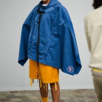 Guest wears orange sunglasses, a blue denim hoodie oversized cloak, yellow orange fringed shorts, yellow socks, white and black gradient print...