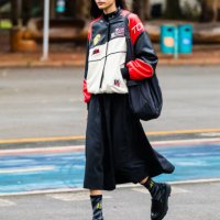 Guest wears racing jacket, black midi skirt, black chunky shoes, black socks with yellow lettering, black oversized bag, black sunglasses at Sao...