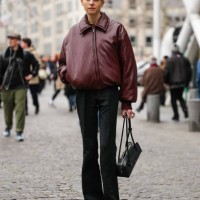 Guest wears shiny burgundy zipped bomber leather jacket, black slim fit flared trouser pants, shiny black Alaïa Le Teckel leather bag, shiny black...