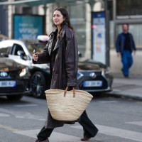 Guest wears shiny dark brown double breasted leather trench coat, light brown basket bag, black relaxed trouser pants, shiny dark brown loafer...
