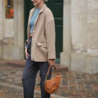 Guest wears silver earrings, a blue gray cropped t-shirt, a beige blazer jacket, gray suit pants, white sneakers, a camel shiny leather Saddle...