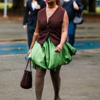 Guest wears sleeveless brown vest, green balloon skirt, beige tights, purple shoes, brown handbag, braided orange hair at Sao Paulo Fashion Week 2024...