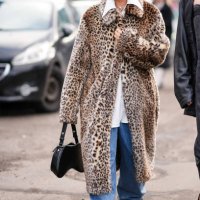 Guest wears sunglasses, a beige leopard print long coat, flared blue denim jeans pants, a black leather bag, outside Skall Studio, during the...