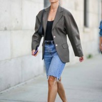 Guest wears sunglasses, a beige oversized blazer jacket, a black low neck top, a belt, blue ripped denim shorts, black pointed flat shoes, outside...