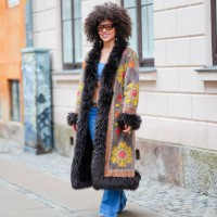 Guest wears sunglasses, a blue bras, a gray and black long winter coat with fluffy parts and printed geometric patterns in yellow and orange, a belt,...