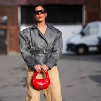 Guest wears sunglasses, a red hair scrunchie designed as a flower, a black leather biker jacket , beige flared pants , a red leather bag outside...