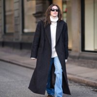 Guest wears sunglasses, a white oversized turtleneck ribbed wool pullover , a black oversized long coat , blue flared denim jeans pants , leopard...