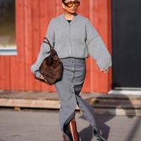 Guest wears sunglasses, earrings, a gray wool oversized cardigan, a brown suede bag, a gray long slit skirt , brown leather boots , outside Gestuz,...