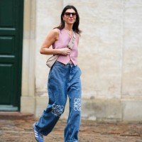 Guest wears sunglasses, gold earrings, gold chain necklaces, a pale pink sleeveless buttoned gilet, a beige Epi leather crossbody bag from Prada,...