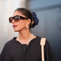 Guest wears sunglasses, golden earrings, a necklace , a scrunchie, a blue monogram logo printed Vuitton bag, a black oversized padded jacket ,...