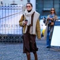 Guest wears sunglasses, golden earrings , a white wool pullover worn as a scarf, a beige aviator jacket with fluffy inner lining , a brown midi...