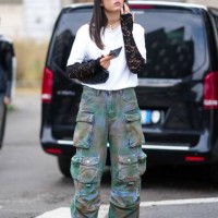 Guest wears sunglasses, necklaces, a white top, black lace mittens, camouflage cargo pants, black leather shoes, outside Anakiki, during the Milan...
