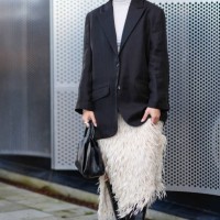 Guest wears sunglasses, silver earrings, a piercing, a white turtleneck, a black oversized blazer jacket, a white fluffy fringed skirt , a black...