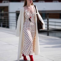Guest wears white knit hat, cream sunglasses, black necklace red pendant, cream midi dress with red floral detail, cream cardigan trench sweater,...