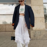 Guest wears white long maxi skirt, white Adidas socks, cropped beige top with green letters and navy blue oversized blazer on day 1 of Visa Fashion...