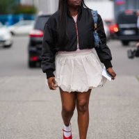 Guest, wears yellow lined glasses with dark brown lens, black cropped bomber jacket, pink shirt, white skirt, white socks, red ballet shoes,outside...
