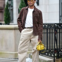 Guest wears yellow sunglasses, a brown suede jacket , a white t-shirt, a belt, beige pants, brown leather pointed shoes, a yellow Loewe bag, outside...