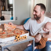 happy man eating pizza while the baby sleeps in his arms - junk food stock pictures, royalty-free photos & images