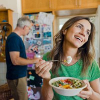 happy mature woman at home eating a salad with man in background - food stock pictures, royalty-free photos & images