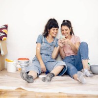 happy twins taking a break while painting at home and using a smartphone - home decoration stock pictures, royalty-free photos & images