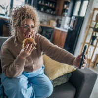 happy woman enjoying a hamburger on the couch while taking a selfie - junk food stock pictures, royalty-free photos & images