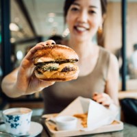 happy young asian woman sitting in a restaurant having her lunch. she is eating cheese burger with fries and a cup of coffee. enjoying a relaxing afternoon - junk food stock pictures, royalty-free photos & images