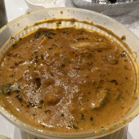 High-angle shot of a plastic carry-out bowl filled with meat and cooked green vegetables in a rich brown gravy, resting on a white tiled surface in...