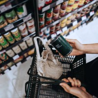 high angle view, close up hands of young asian woman grocery shopping in supermarket. she is putting a tin can into a cotton mesh eco bag in a shopping basket. environmentally friendly and zero waste concept - food fotografía