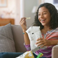 hispanic girl eating potato chips on sofa - junk food stock pictures, royalty-free photos & images