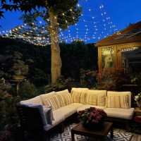 image de la zone de détente extérieure la nuit éclairée par des lumières de fées à cordes, des sièges en bois franc avec coussins, une table en bois avec une pièce maîtresse de plante à fleurs, des bonsaïs, des érables japona